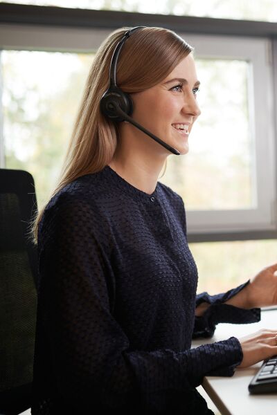 A woman sits at her desk, wearing a headset, typing on her keyboard with a smile infront of two displays with SIXT slides in orange..