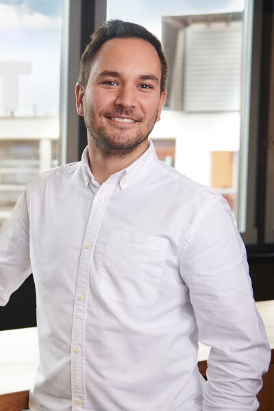 A smiling man stands at a table in his office, holding a pen and a notepad. Behind him, a large window offers a view of the SIXT office, prominently displaying the SIXT logo.