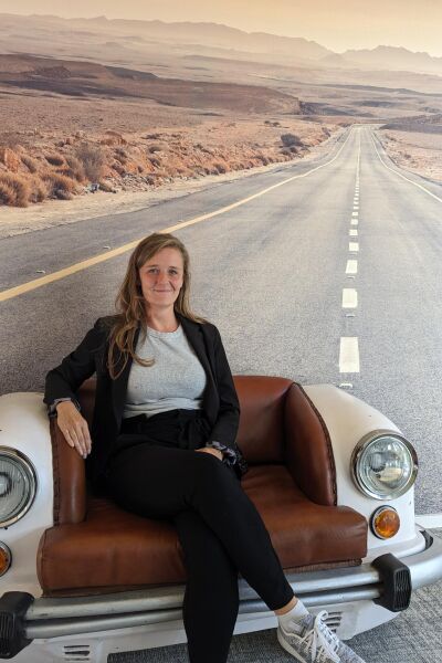 Mona sits in an armchair designed to resemble the rear of a car, positioned in front of an image that depicts a long road stretching into the distance. She is smiling.