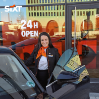 A rental sales agent in her uniform is standing smiling next to a car in front of a Sixt branch with the sign: "24h open".