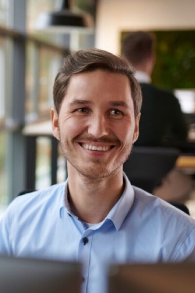 A man sits in front of his screens, laughing, while another man works at his desk in the background.