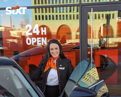 A rental sales agent in her uniform is standing smiling next to a car in front of a Sixt branch with the sign: "24h open".