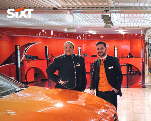 Two Rental Sales Agents in uniform standing in a branch behind an orange car, smiling warmly.