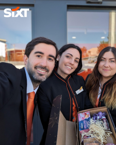 Three rental sales agents in uniform standing in front of a SIXT branch, holding an Easter basket while they are smiling.