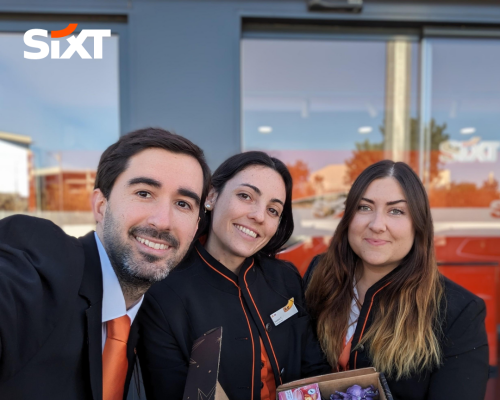 Three rental sales agents in uniform standing in front of a SIXT branch, holding an Easter basket while they are smiling.