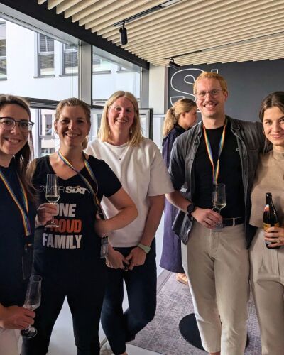 Five people stand in an office, holding drinks and a keychain in pride colors while smiling.