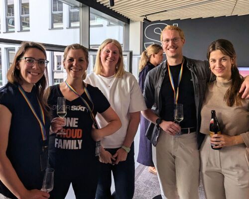 Five people stand in an office, holding drinks and a keychain in pride colors while smiling.