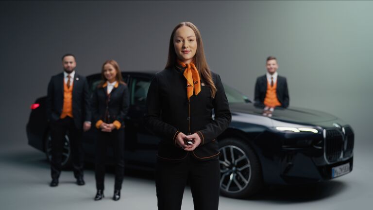 In the foreground, a woman dressed in the SIXT uniform holds a set of car keys, while in the background, a BMW is visible along with three other Sixt employees.