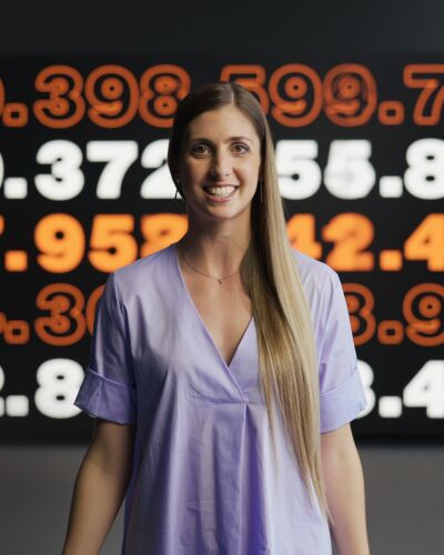 A woman is smilling. In the background, there's a board with various numbers on it.