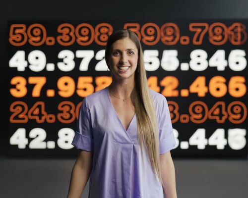 A woman is smilling. In the background, there's a board with various numbers on it.