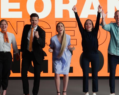 A group of people in various outfits is cheering together. In the background, there’s a board in SIXT colors with the word "Orange" written on it.