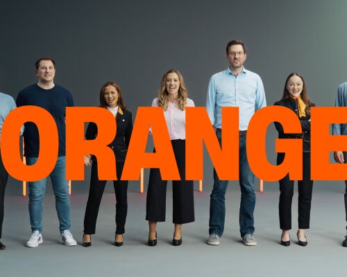 A group of people standing in a row, each with different facial expressions, with the word "ORANGE" displayed in front of them.
