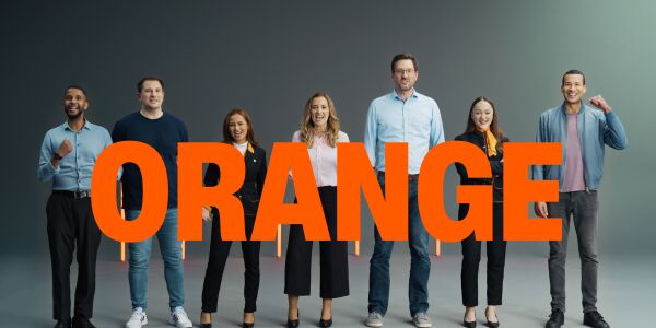 A group of people standing in a row, each with different facial expressions, with the word "ORANGE" displayed in front of them.