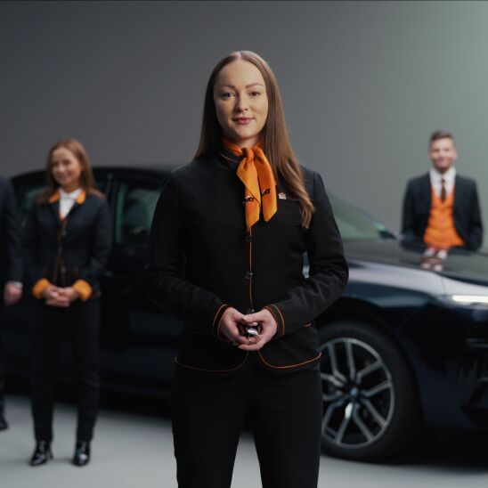 In the foreground, a woman dressed in the SIXT uniform holds a set of car keys, while in the background, a BMW is visible along with three other Sixt employees.