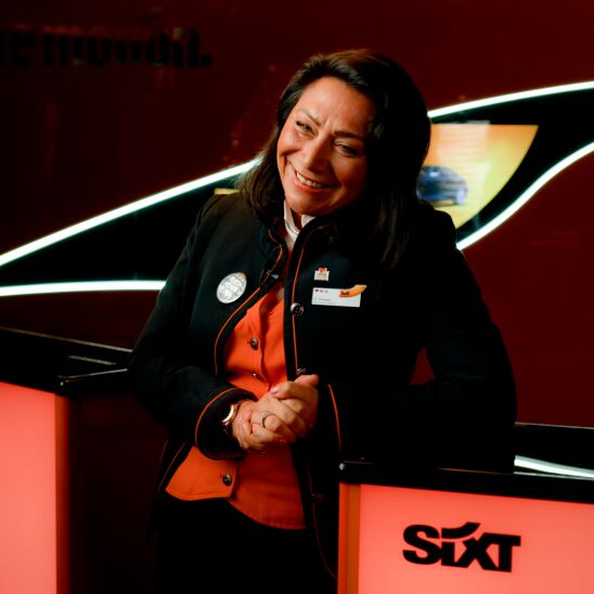 A Rental Sales Agent in her SIXT uniform stands smiling in a SIXT branch.