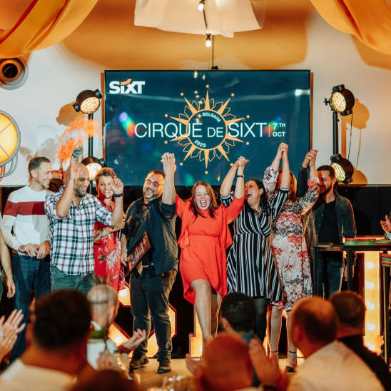 People standing on stage, cheering, with a screen behind them displaying the words "Cirque de SIXT." In front of them is a seated audience, applauding.